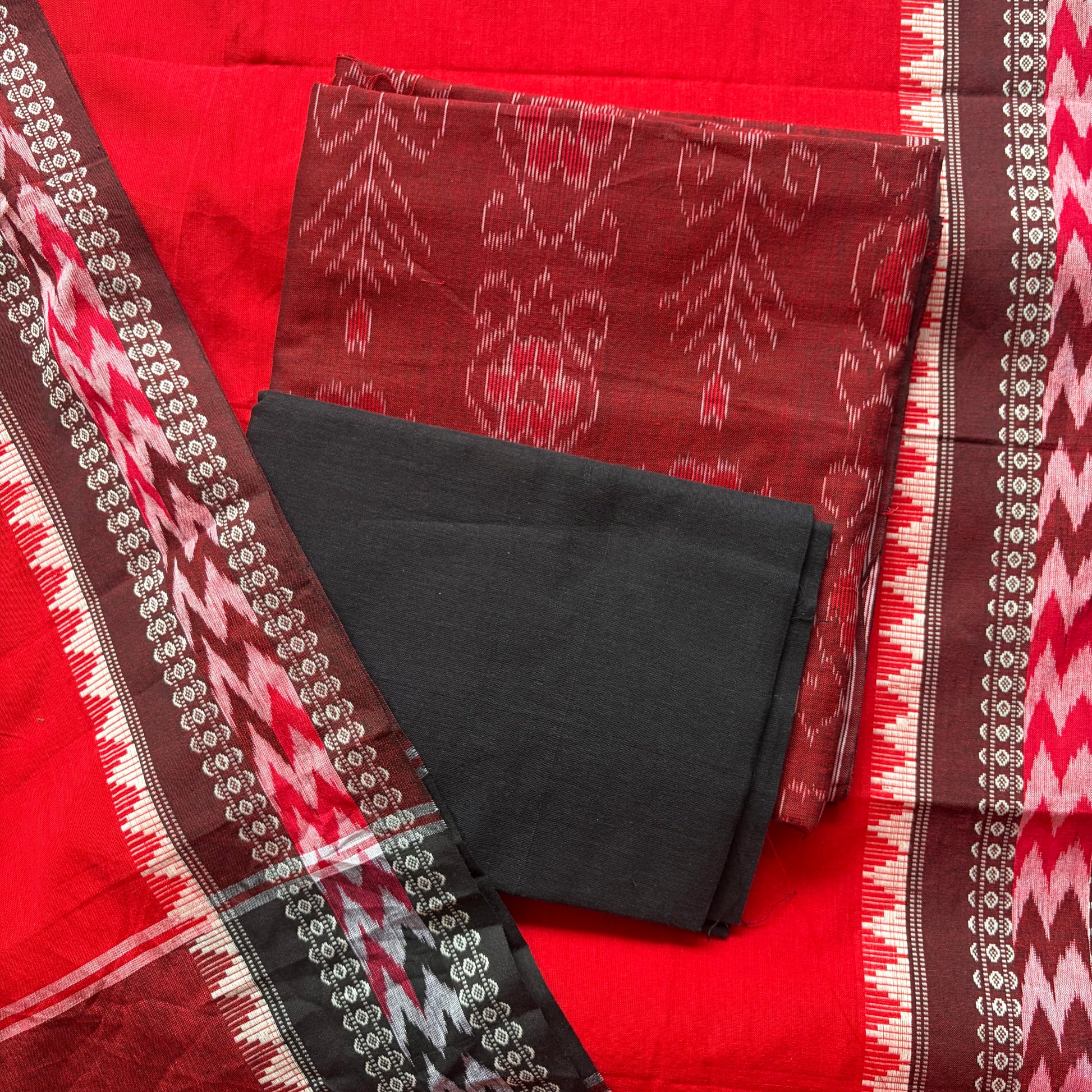 A folded traditional Indian saree fabric set featuring a rich red body, a dark reddish-brown patterned pallu, and a solid black fabric piece, with decorative borders showing red, white, and brown ikat and zigzag motifs.